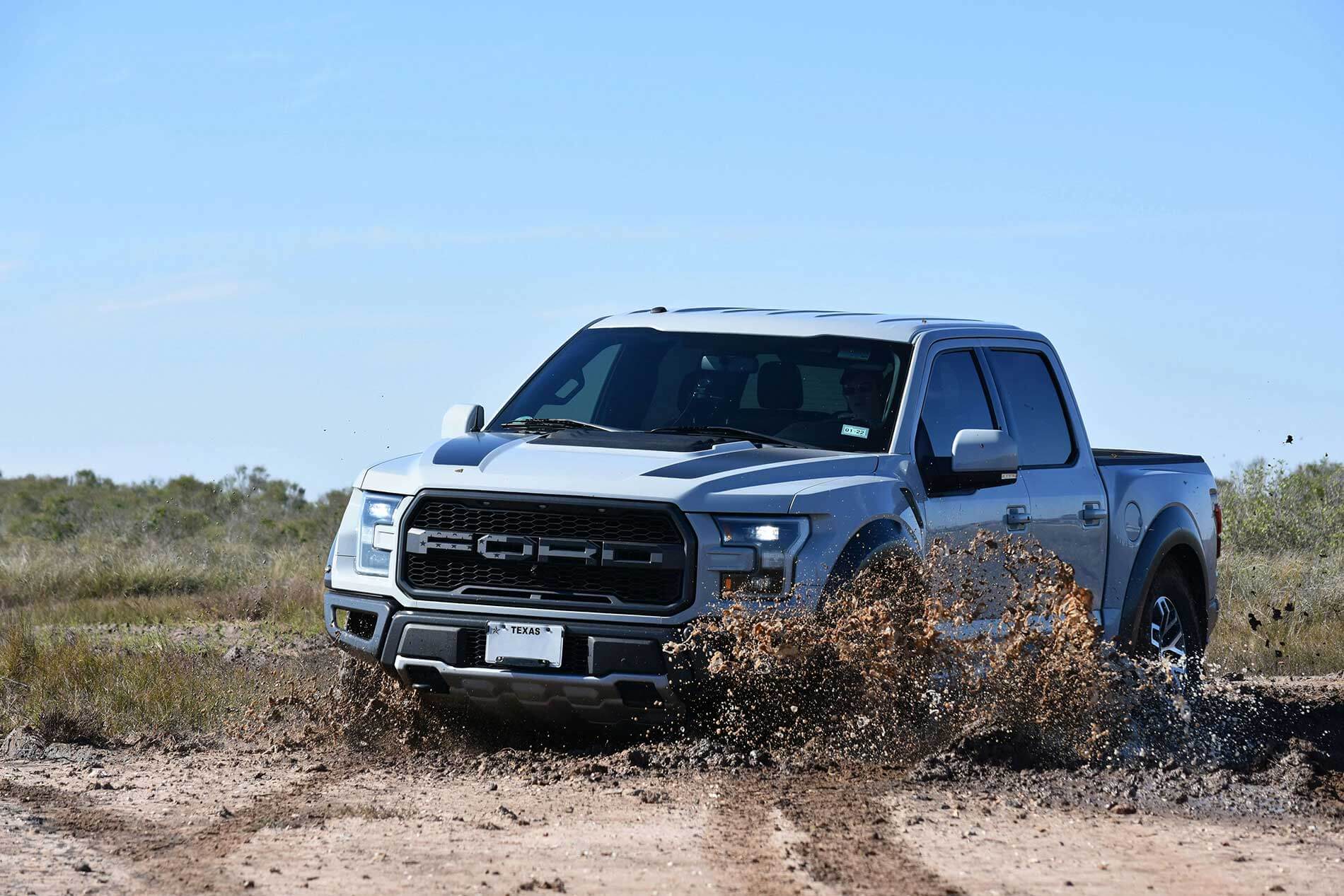 A silver Ford F-150 Raptor pickup truck driving through mud in a rural Texas landscape. The truck is kicking up a large spray of mud and dirt as it moves through rough terrain. The vehicle has a Texas license plate and is set against a background of scrubland and clear blue sky.