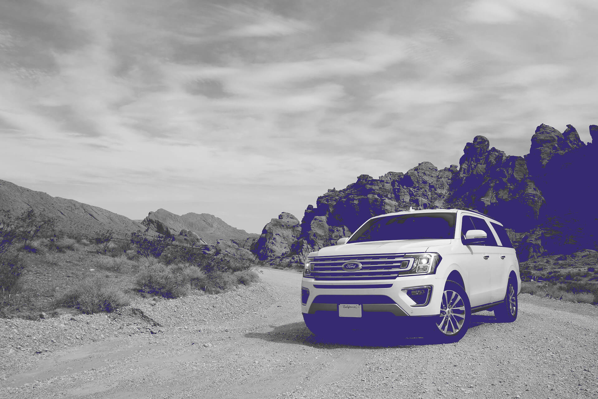 A white Ford SUV parked on a dirt road in a desert landscape. The vehicle is surrounded by rugged red rock formations and mountains under a partly cloudy blue sky. Sparse desert vegetation can be seen in the foreground. The scene appears to be in a place like the American Southwest, possibly a national park or scenic area.