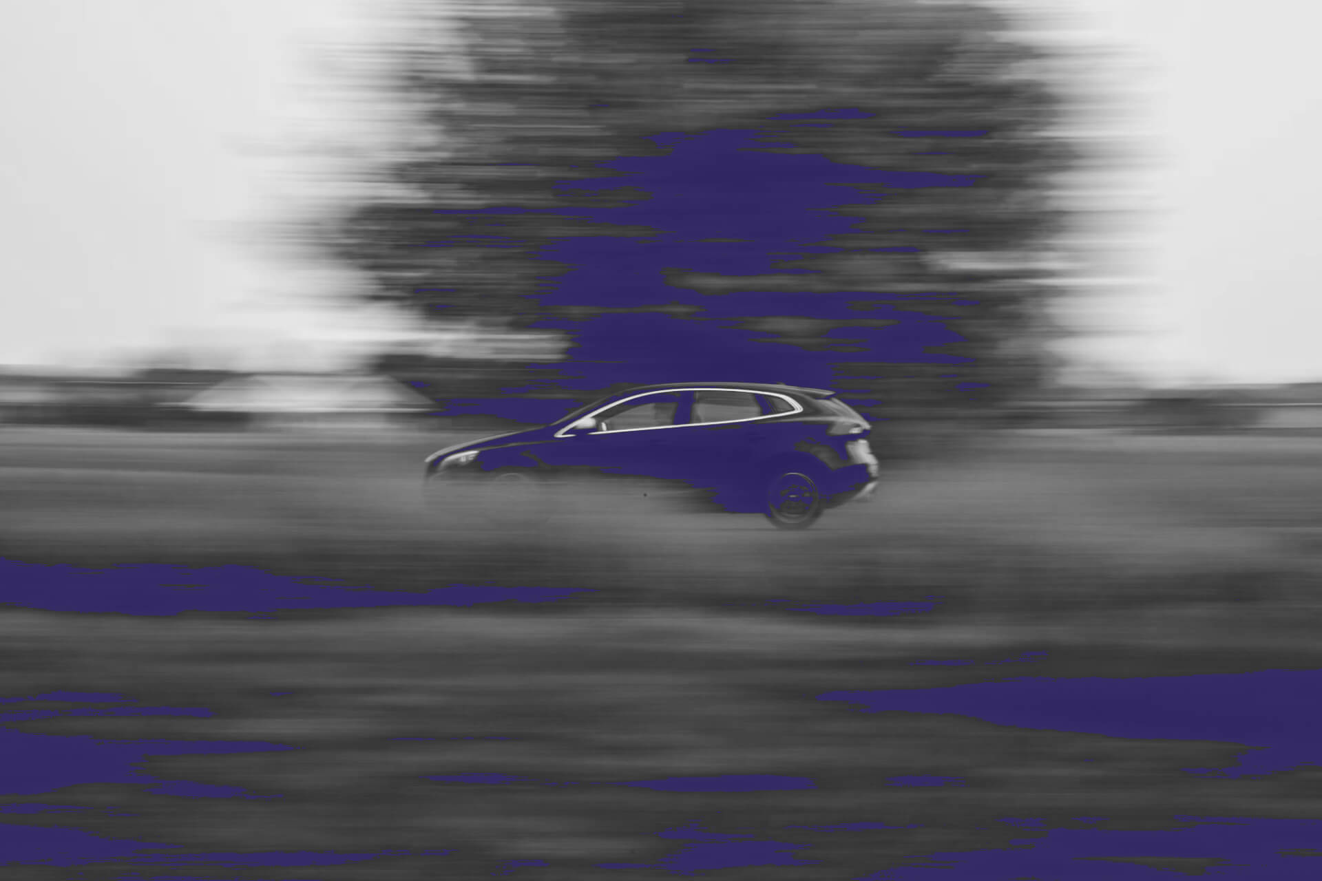 A blurred black and white image of a car driving on a road, with purple color accents highlighting the vehicle and parts of the background. The car appears to be a hatchback or small SUV in motion, creating a sense of speed. The surrounding landscape is indistinct, with a large blurred shape, possibly a tree, visible behind the car. Purple patches are scattered across the foreground, possibly representing grass or flowers.